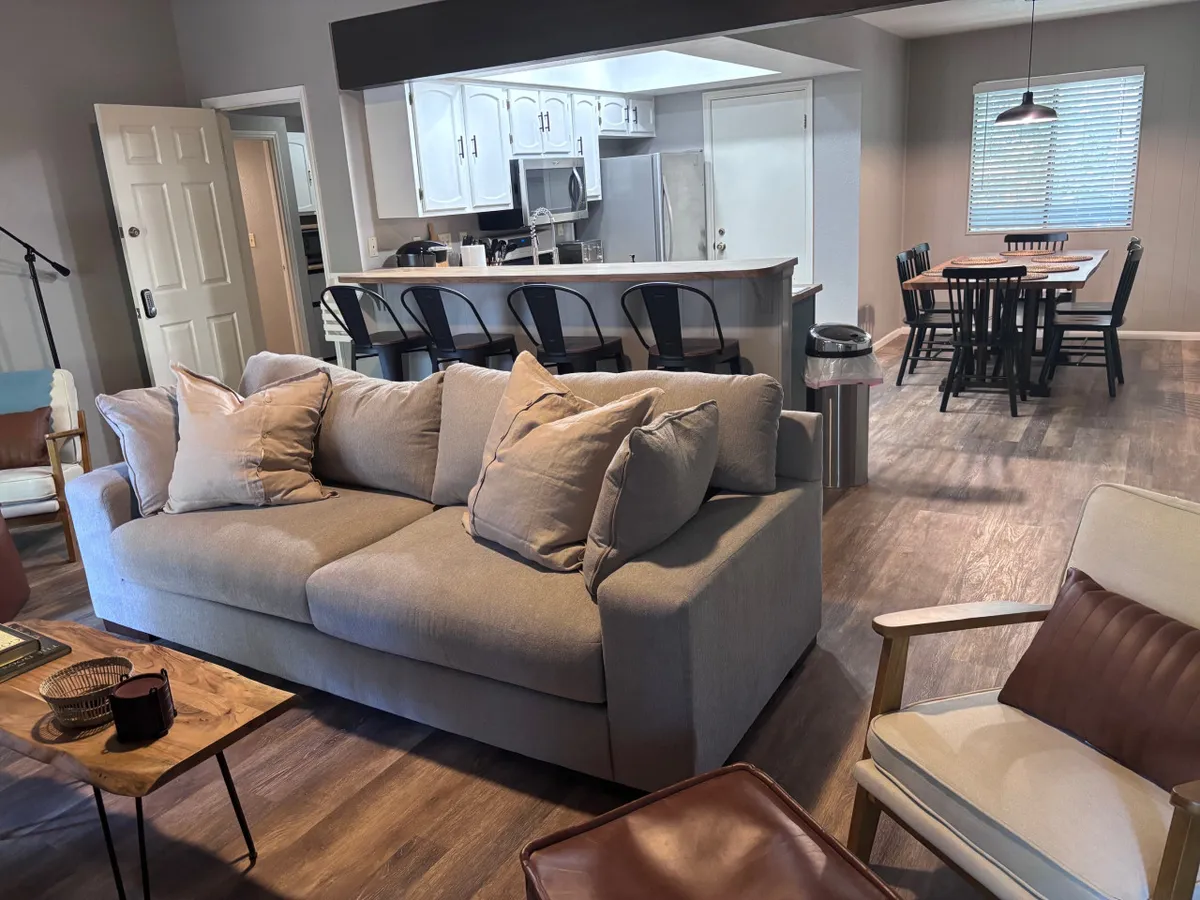 This inviting scene captures a plush sofa accented with pillows, a spacious dining table, and a modern kitchen bar area in the background. Natural sunlight streams into the open-plan space, highlighting the seamless flow between relaxing, dining, and cooking zones. The overall vibe is cozy yet lively, perfectly suited for gatherings and shared moments.