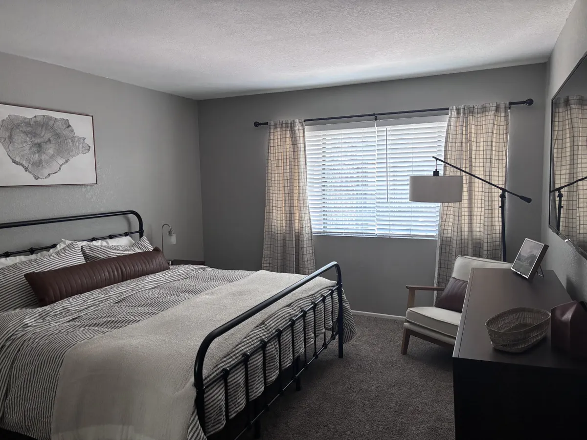 This image displays a serene bedroom with a bed featuring striped bedding, bathed in natural light from a large window. A modern lamp and decorative wall art add to the tranquil and cozy ambiance, inviting relaxation and comfort.