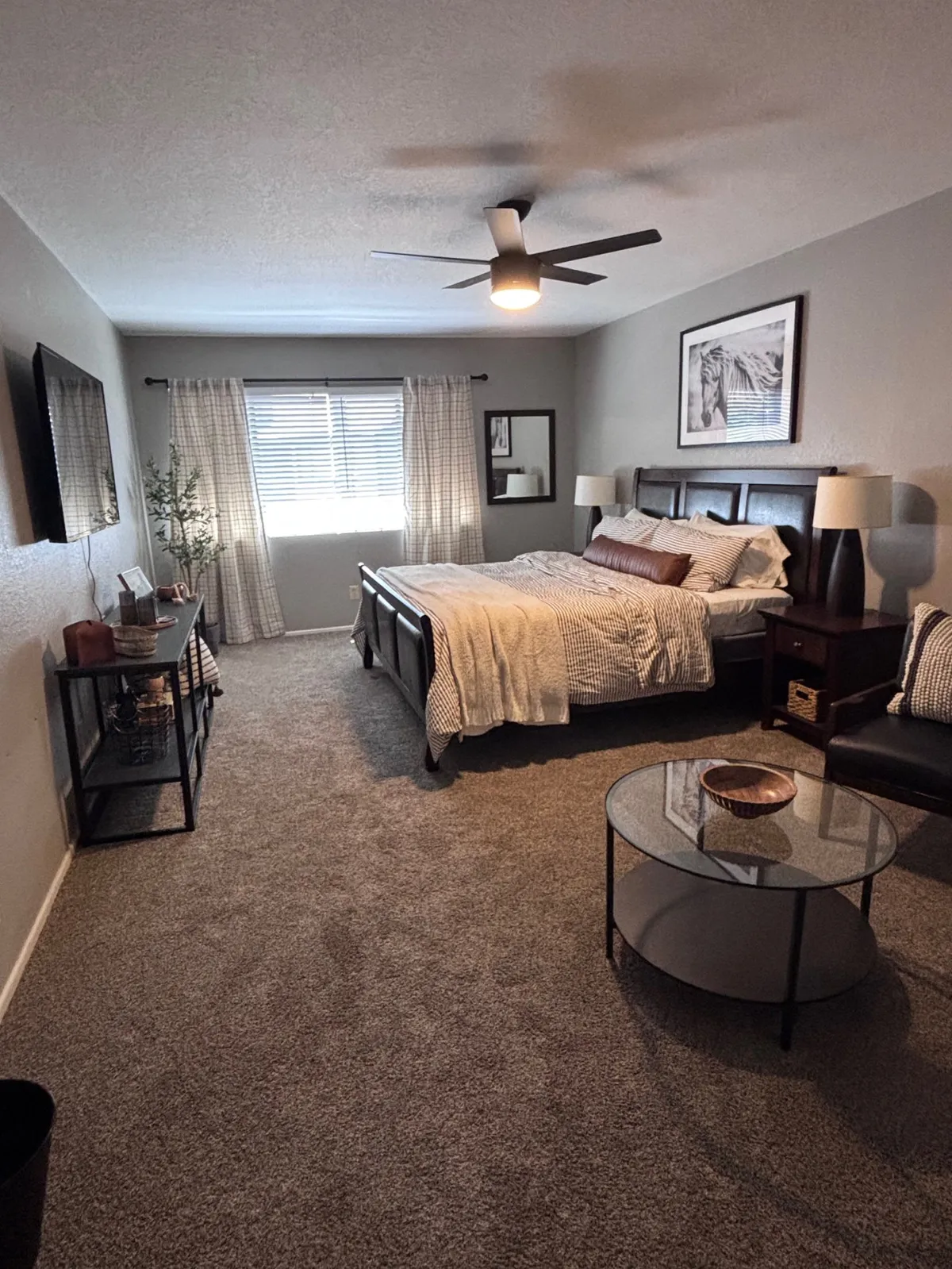 This image features a cozy bed with decorative pillows, a modern ceiling fan, stylish nightstands, and artwork. The large window with curtains allows for natural light, creating a soothing, inviting atmosphere perfect for relaxation and quiet moments.