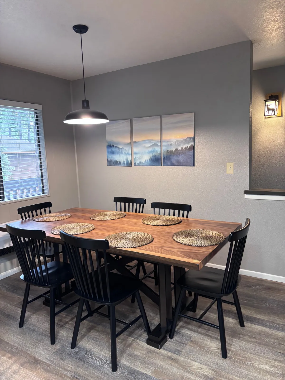 A cozy, welcoming dining area featuring a large table surrounded by black chairs. Wall art adds a homey touch, and the space is clearly designed for group gatherings, family meals, or celebrations. The natural light streaming in hints at a bright, inviting atmosphere.