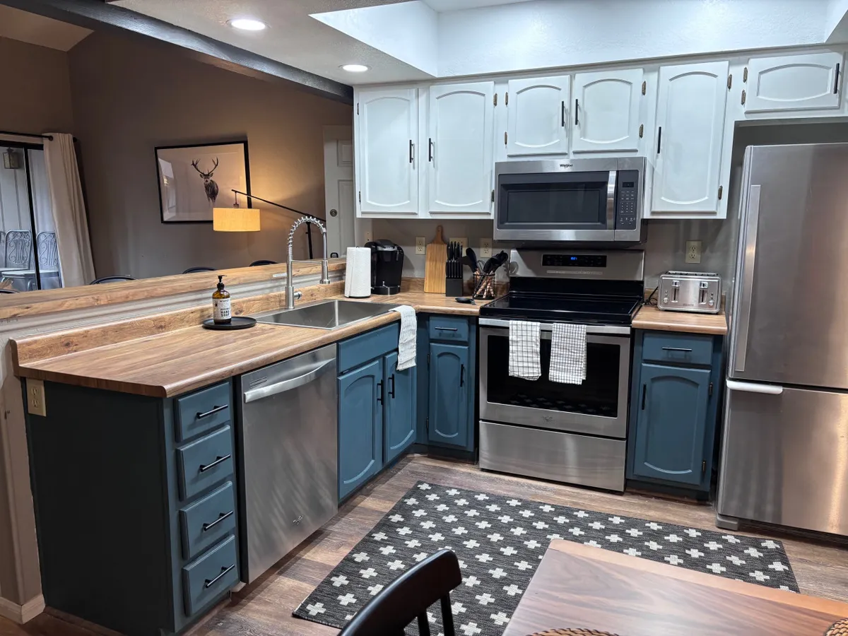The image showcases a beautifully remodeled kitchen and great room area, featuring a large wooden island with seating, sleek stainless steel appliances, and a stylish patterned rug. The space feels open and inviting, perfect for family meals or entertaining guests against a backdrop of cozy mountain elegance.